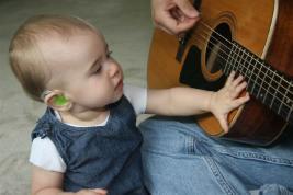 Toddler wearing an assistive hearing device looking at a person with a guitar